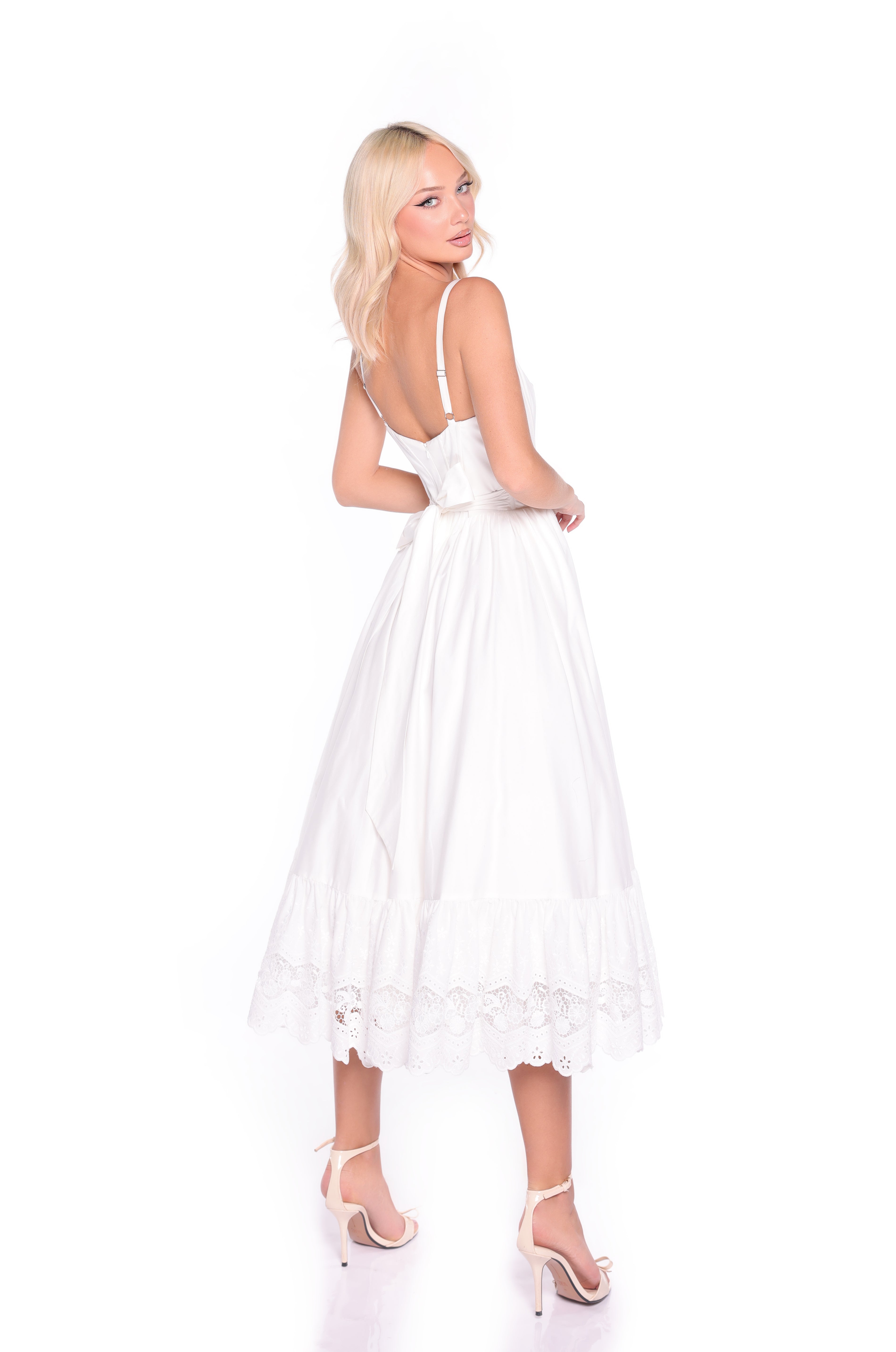 A woman with blonde hair wears the White Casual Midi Dress: Celeste—sleeveless, with lace trim and a corset bodice—paired with white high-heeled sandals, posed against a plain white background.